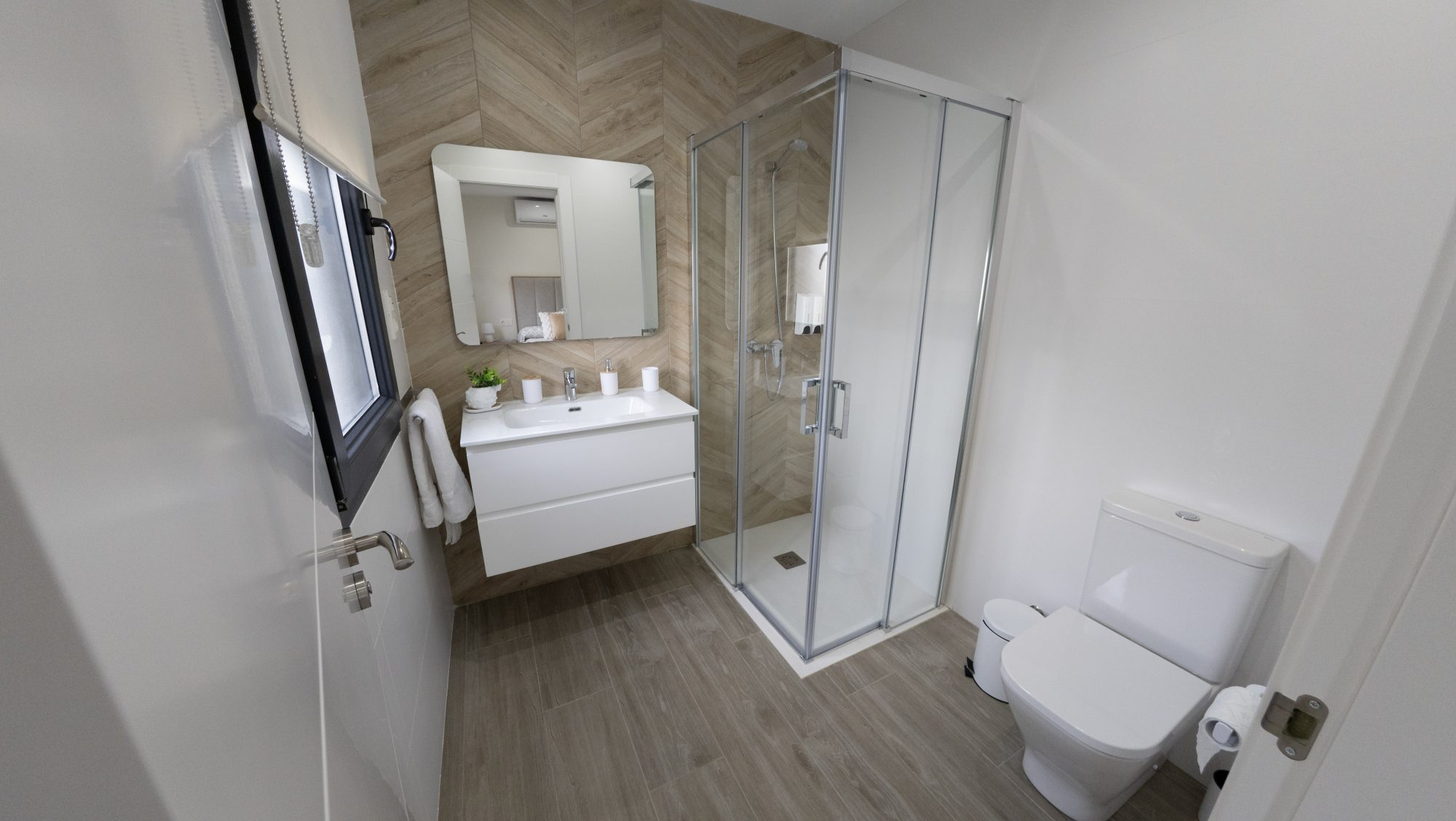 Baño con espiga de madera, mueble blanco y ducha de cristal.
