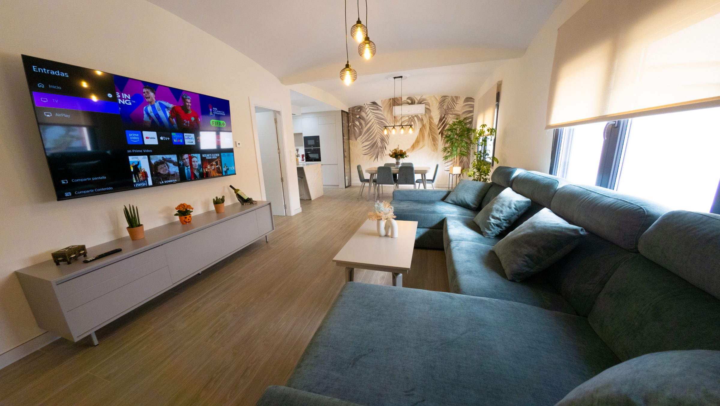 Salón con sofá modular azul, televisión de gran formato y comedor al fondo bajo lámparas colgantes.