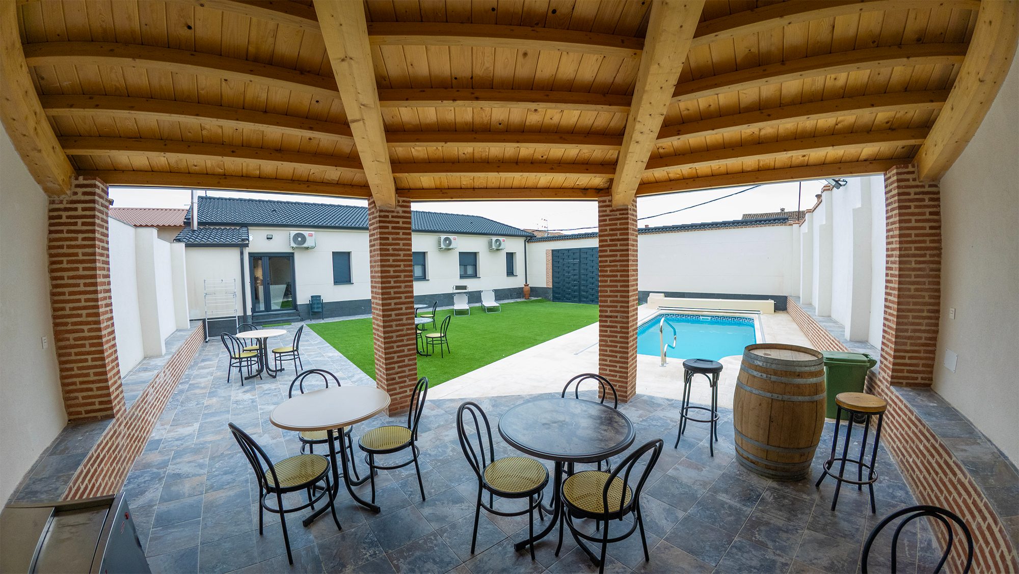 Merendero cubierto con vigas de madera, mesas y sillas frente a una piscina privada y césped.